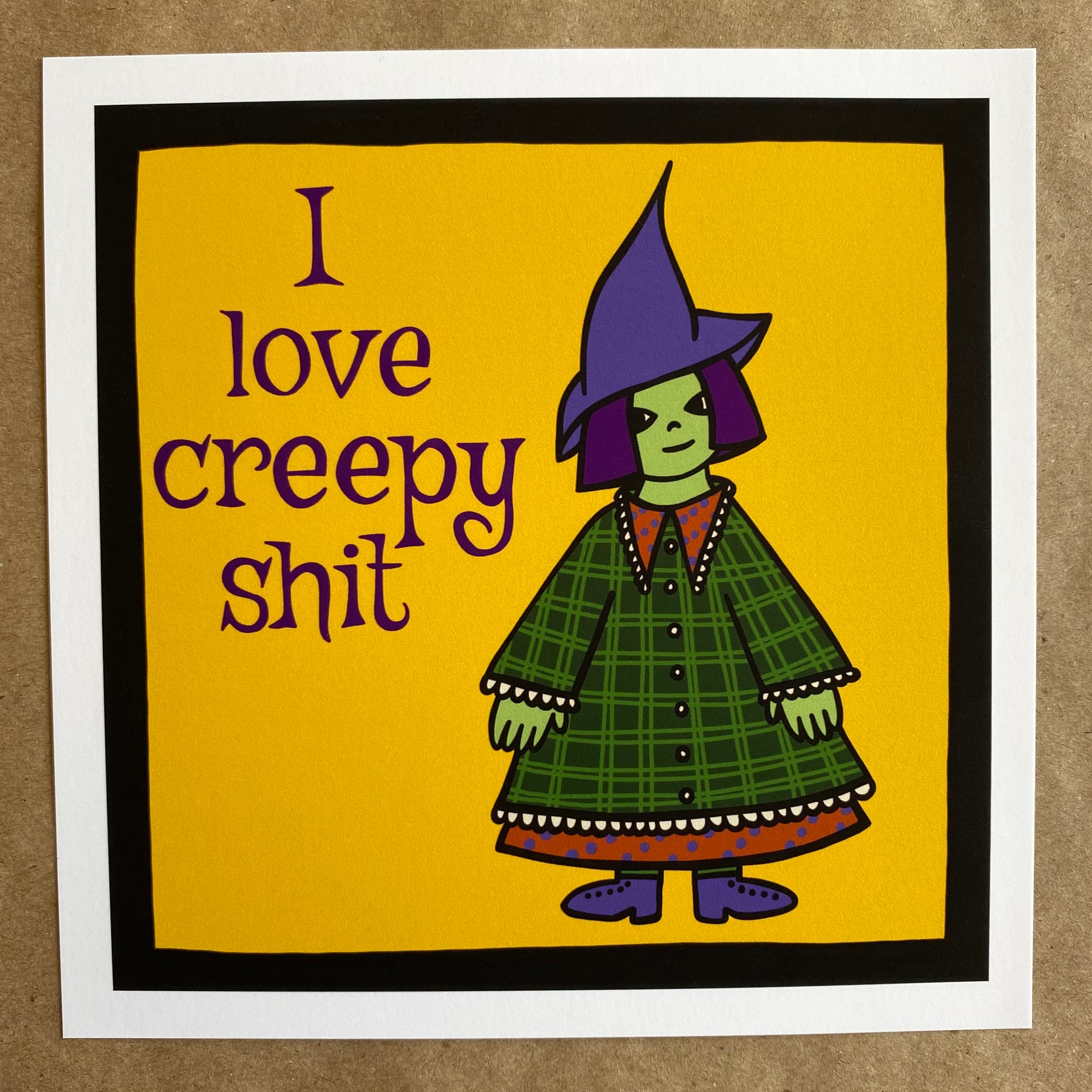 a photo of an art print featuring original art by AnneArchy, of a green-skinned witch wearing a green checked coat over an orange dress with purple polka dots; with hand lettering of the words: I love creepy shit