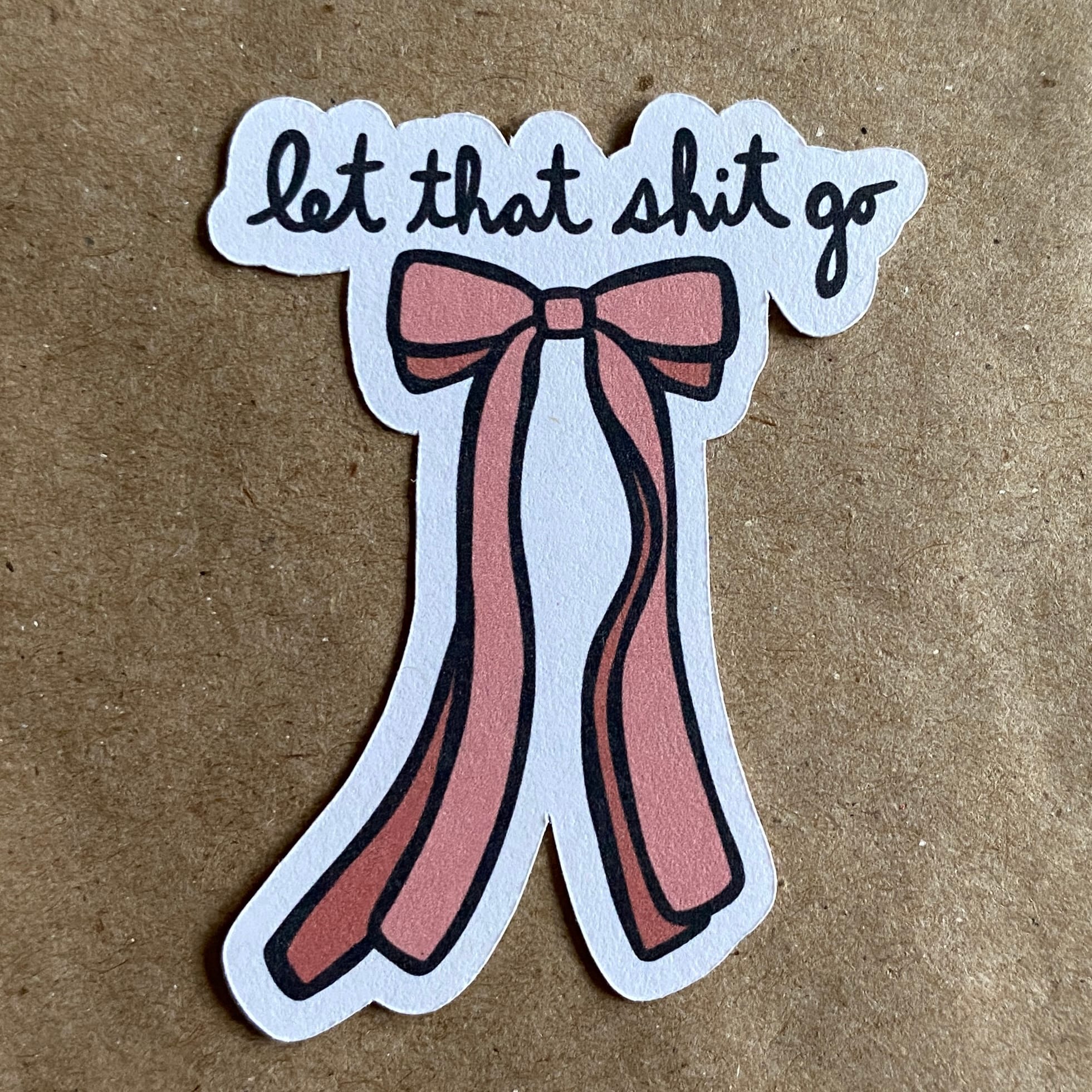 a photo of an original art sticker by AnneArchy featuring a drawing of a pink ribbon with hand lettering of the words "let that shit go"