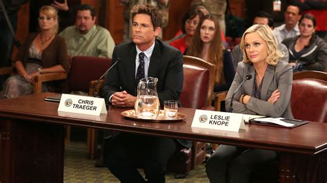 a still from the TV show Parks and Recreation, in which Rob Lowe as Chris Traeger and Amy Poehler as Leslie Knope are seated at a table behind microphones and name signs