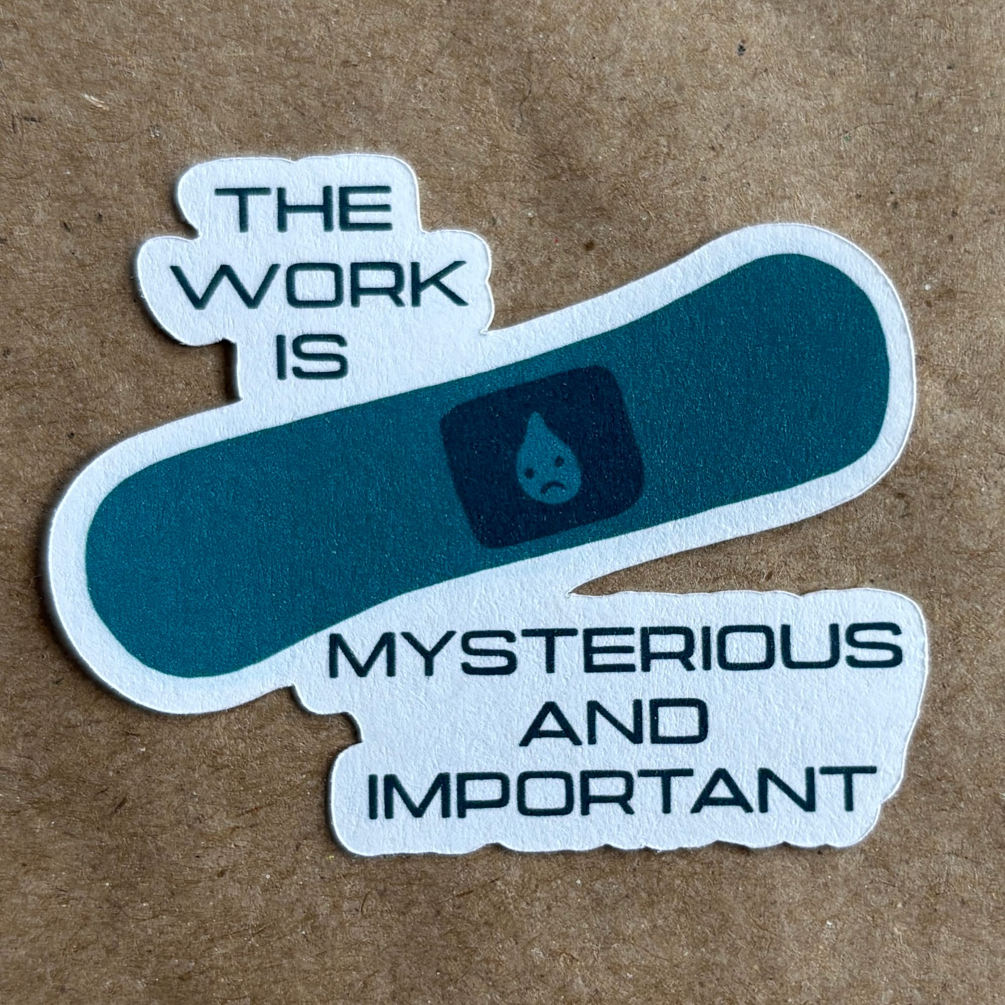 a photo of an original art sticker by AnneArchy featuring a drawing of a Lumon sad-face bandage with hand lettering of the words "the work is mysterious and important"